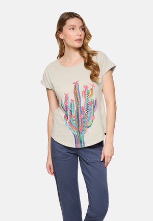 Mujer con camiseta beige con un gráfico colorido de cactus estampado y pantalones casuales azules, de pie con la cabeza girada hacia la derecha.