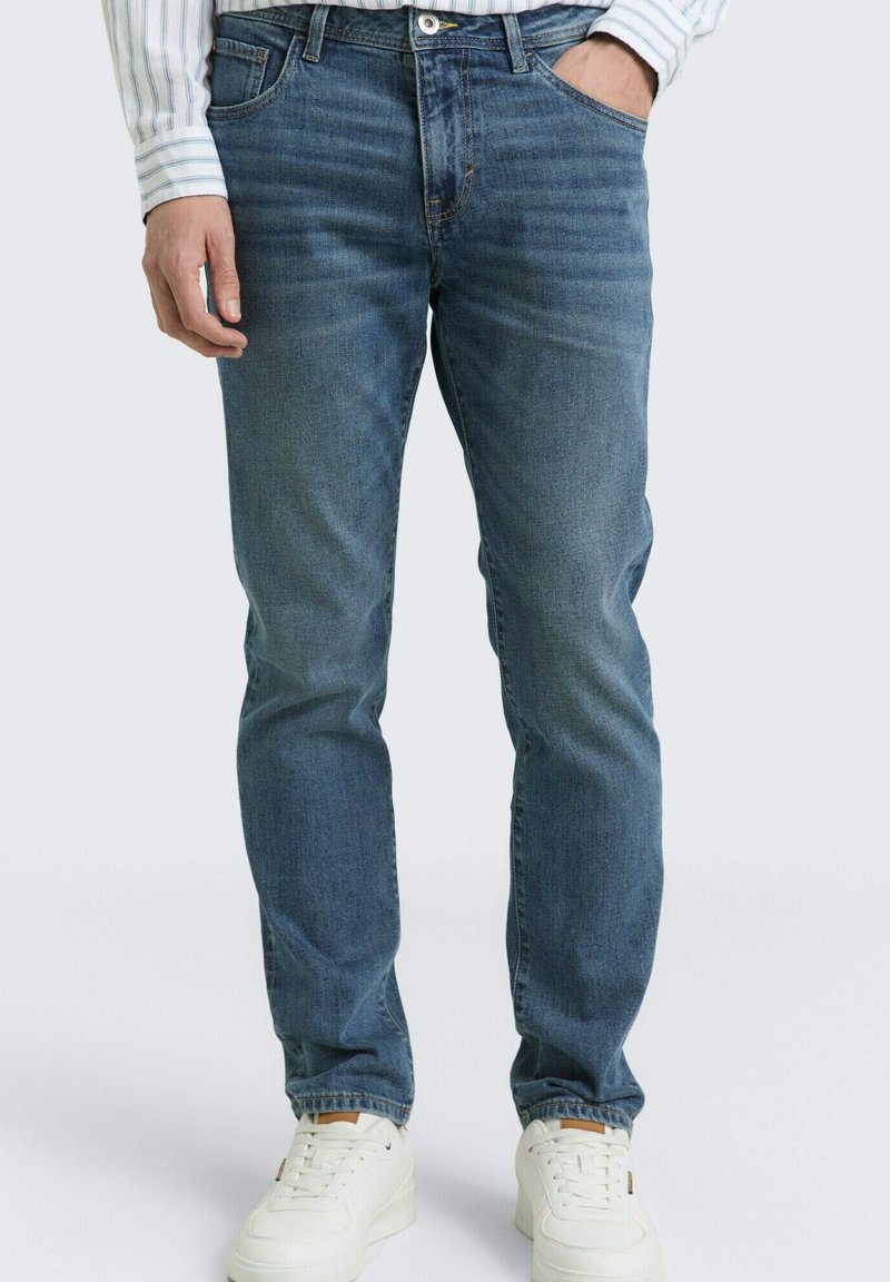 Man draagt blauwe slim-fit jeans, witte sneakers en een gestreept shirt met lange mouwen, met één hand in de zak.