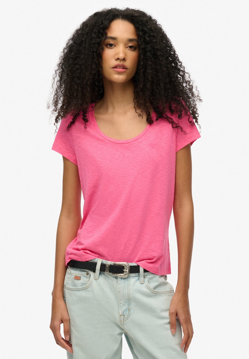 Frau mit lockigem Haar, die ein hellrosa T-Shirt mit weitem Ausschnitt trägt, das leicht in eine hellblaue Jeans mit schwarzem Gürtel gesteckt ist.