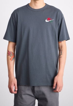 Homme portant un t-shirt gris foncé avec un petit swoosh blanc et un motif de rose rouge sur le côté gauche de la poitrine, tatouages colorés visibles sur les deux avant-bras.