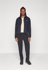 Marineblaue Fleece-Jacke mit Reißverschluss, ausgestattet mit zwei Vordertaschen. Kombiniert mit schwarzen Hosen, die mit Reißverschlussakzenten versehen sind, und robusten schwarzen Stiefeln.