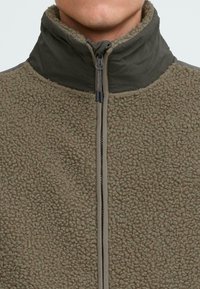 Giacca in fleece di colore verde oliva con superficie testurizzata, colletto alto e chiusura frontale con zip; tessuto liscio a contrasto sul colletto e sulle spalle.