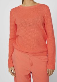 Korallfärgad stickad tröja med ett texturerat mönster, rund halsringning och ribbade ärmslut. Matchas med korallfärgade byxor.