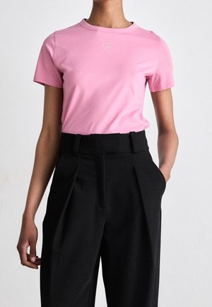 Person iført en tætsiddende pink t-shirt med et lille hjerte-design og højtaljede sorte plisserede bukser med hænderne i lommerne.
