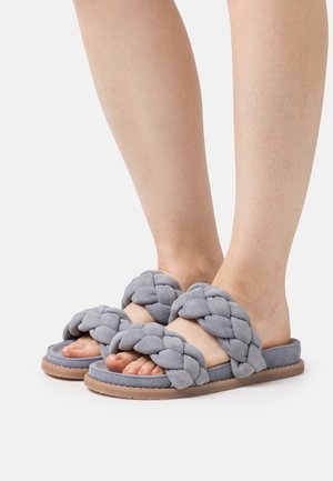 Piernas con sandalias sin talón de tiras trenzadas grises y suelas planas de color marrón claro sobre un fondo blanco liso.