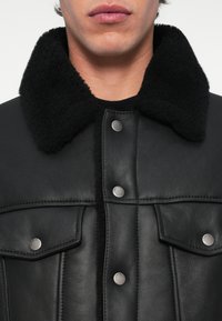 Casaco de cabedal preto com um colarinho de forro felpudo preto texturizado, botões de pressão prateados e bolsos frontais. Acabamento suave com design estruturado.