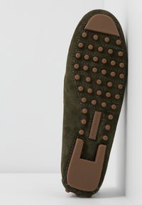Semelle de chaussure en daim vert avec des grips en caoutchouc marron et étiquette "TORY BURCH" affichée verticalement sur fond blanc.