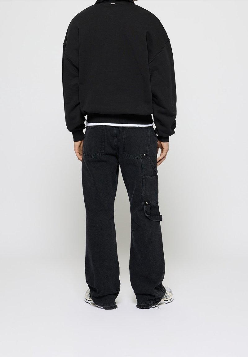 Personne portant un pantalon cargo noir ample et un sweat-shirt noir, debout, de dos, sur un fond clair uni.