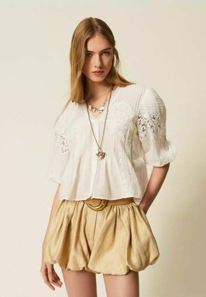 Joven mujer lleva blusa blanca de encaje con mangas abullonadas y pantalones cortos beige texturizados, complementados con collares largos con colgantes y un cinturón.