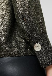 Blusa nera con design a pois dorati, caratterizzata da un accento di bottone rotondo in argento sul polsino e tessuto nero liscio sull'orlo.