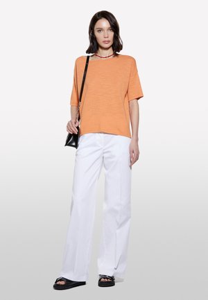 Haut orange à manches courtes en tissu léger, associé à un pantalon large blanc. Un sac bandoulière noir et des sandales plates complètent le look.
