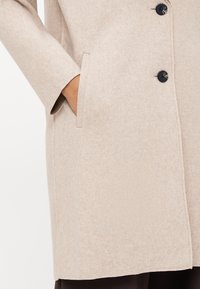 Beige wolmix jas met een rechte silhouet, voorzien van twee zijzakken en vier zwarte knopen aan de voorkant. Zachte, gestructureerde stof.