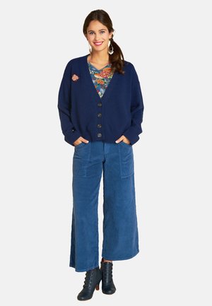 Marinefarbener Cardigan mit großen Knöpfen, kombiniert mit blauen weiten Cordhosen und schwarzen Absatzstiefeln. Blumenshirt darunter sichtbar.