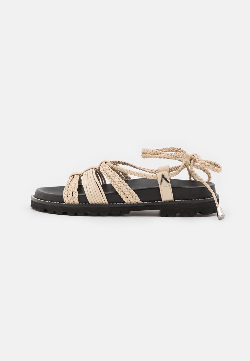 Sandales beige tressées avec une semelle noire, présentant un design plat, des liens à la cheville, et un motif texturé sur les lanières.