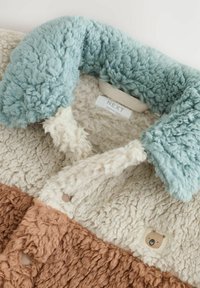 Veste en polaire texturée avec un col bleu clair, des sections crème et marron, et un petit patch d'ours près de l'avant. Étiquette visible à l'intérieur.