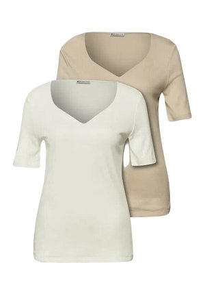 Kvinde med kort blond hår iført en beige kortærmet skjorte og lyseblå jeans, set fra siden med en neutral baggrund.