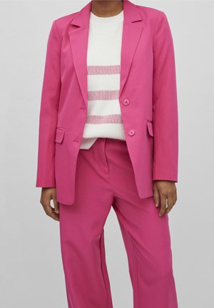 Person trägt einen leuchtend pinken Blazer und passende Hose über einem weißen Pullover mit horizontalen pinken Streifen, steht vor einem schlichten Hintergrund.
