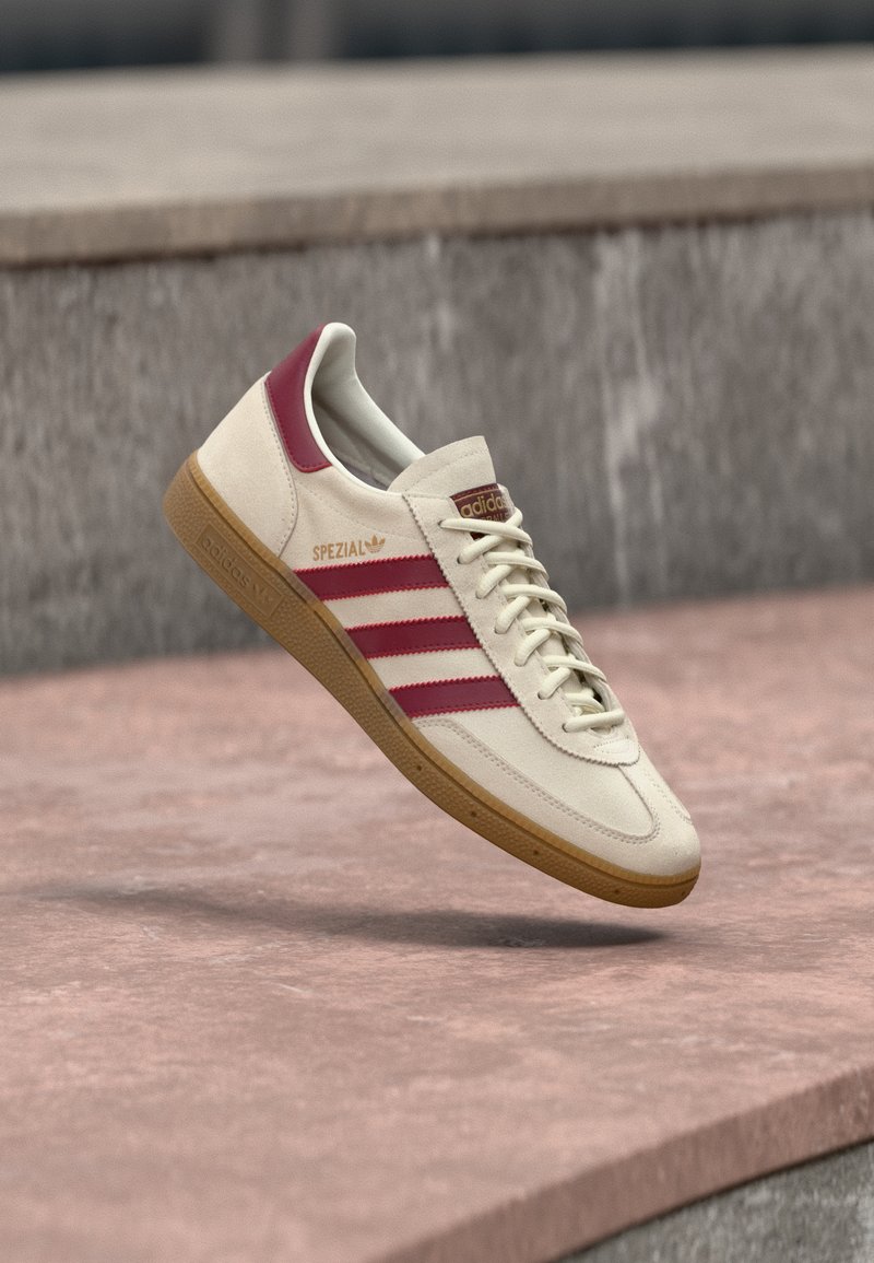 Beige Adidas Spezial-sneaker med mørkerøde striber og hælfane, tanfarvet gummisål, svævende over en pink og grå betongulv.
