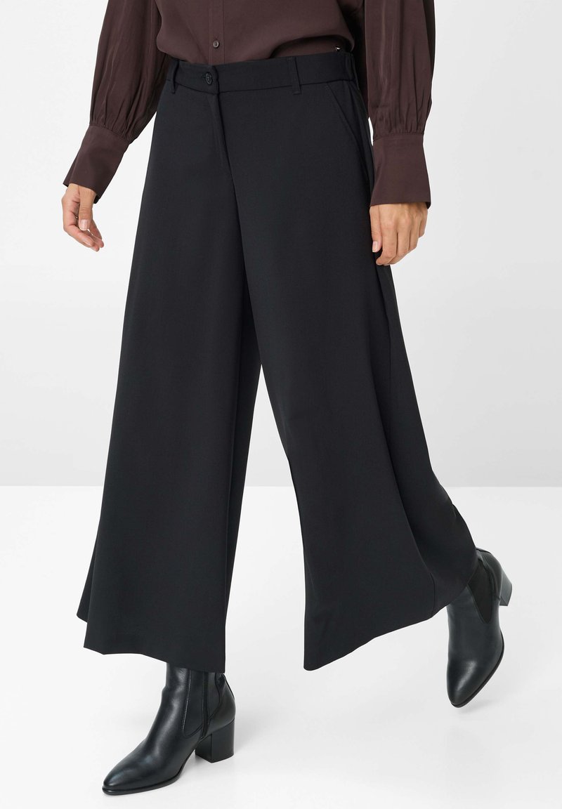 Pantalons noirs à jambes larges en tissu lisse, avec une fermeture à bouton et des poches latérales, associés à des bottines noires.