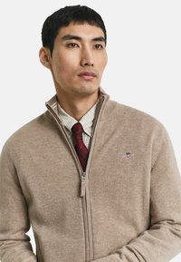 Man in beige ritsvest over een geruite shirt en een rood geruit strikje, iets naar rechts kijkend op een effen achtergrond.