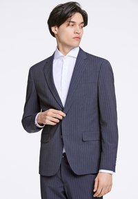 Mörk rutig kostym med nakklaffar, matchande byxor och en vit skjorta. Har två framfickor och knappdetaljer.