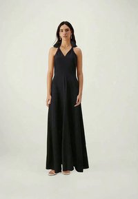 Naine kannab õlavarteta musta halterneck maksimek ootamas avatud varvastega valgeid sussikesi, seismas ühetaolise heleda tausta ees.