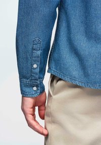 Chemise en denim bleu clair avec poignets boutonnés. Tissu texturé avec des détails de couture visibles. Main reposant contre un pantalon beige sur le côté.