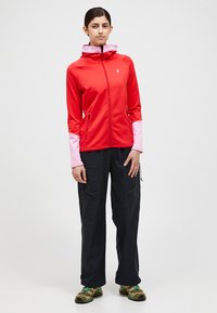 Red zip-up jacket with pink sleeves and hood trim, featuring side pockets and a logo. Worn with black cargo pants and multi-colored shoes.