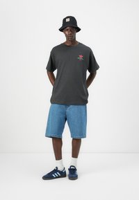 Cappello a secchio nero, t-shirt in cotone grigia con grafica rossa e verde, pantaloni corti di jeans blu e sneaker navy con strisce azzurre.