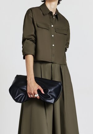 Person trägt olivgrünes, knöpfbares Hemd und plissierten Rock, hält große schwarze gefaltete Leder-Clutch mit silbernen Ringen in der Hand.