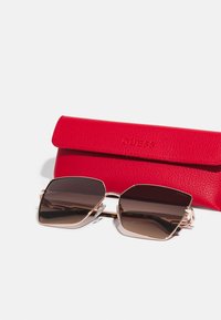 Ochelari de soare hexagonali cu lentile maro cu degradé și rame subțiri aurii rose, însoțiți de o carcasă texturată roșie inscripționată "GUESS."