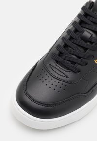 Černé kožené tenisky s hladkým povrchem, kulatou špičkou a perforacemi. Obsahují černé tkaničky a kontrastní bílou gumovou podrážku.