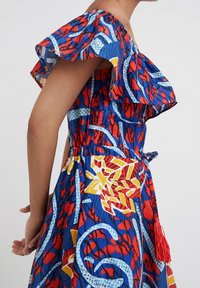 Vrouw in een mouwloze, off-shoulder jurk met abstracte patronen in rood, blauw, wit en geel, en een rode kwastbroek, zijaanzicht.
