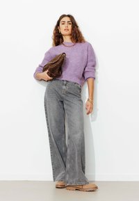 Femme en pull lilas et large jean gris tenant un sac en imprimé léopard, portant des chaussures beiges et des bijoux en or sur fond blanc.