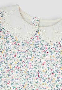 Weißer Stoff mit einem Blumenmuster in Rosa, Gelb und Blau. Mit einem gewellten, mit Augenbrauen versehenen Kragen und einem Knopfverschluss.