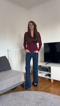 Bordeauxroter Strickpullover mit V-Ausschnitt, kombiniert mit weitem, dunkelblauem Jeansstoff. Lange, glatte Haare. Schwarze Hausschuhe auf einem Holzboden.