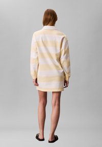 Femme debout, dos tourné, portant une robe-chemise de rugby à manches longues et ample, avec de larges rayures crème et jaune pâle, et des sandales noires à enfiler.