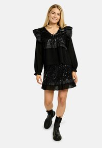 Melna kleita ar spīdumiem, volānu kakla izgriezums un piedurknes, kārtots svārku virsējais slānis ar volānu, valkāta kopā ar melnām potīšu zābakiem. Gluda un teksturēta auduma.