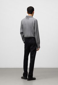 Camicia grigia a maniche lunghe con una vestibilità rilassata e colletto, caratterizzata da un discreto dettaglio del logo sul retro, abbinata a pantaloni neri e calzature.