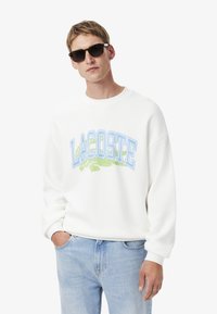 Felpa bianca con logo "LACOSTE" blu e verde, vestibilità ampia, scollatura rotonda, indossata con jeans in denim chiaro e occhiali da sole neri.