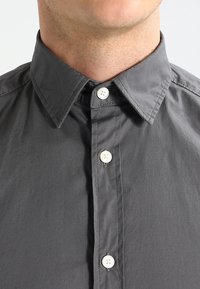 Grå skjorte med knapper, klassisk krave, hvide knapper og glat stofstruktur. Synlige syningsdetaljer langs knaplisten.