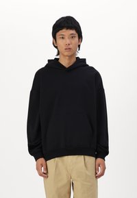 Schwarzer Kapuzenpullover aus weichem Stoff, mit einer vorderen Tasche und elastischen Bündchen, getragen mit hellen Hosen.