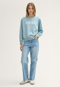 Light blue sweatshirt with "AMORE" text, crew neck, and ribbed cuffs, paired with light blue denim jeans and white sneakers.