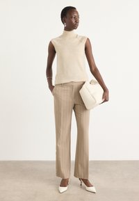 Bež brezrokavnik z visokim ovratnikom, tan prugaste hlače, bele visoke pete in bela torbica. Gladke teksture in kroj po meri.