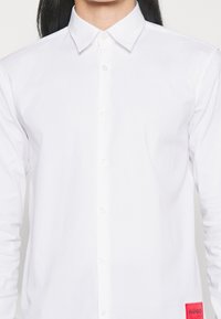 Vit långärmad skjorta i bomull. Har en knapp ner-krage, framknäppning och en röd märkespatch vid nederkanten.
