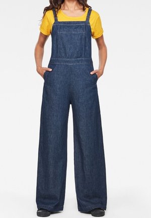 Ogrodniczki z denimu o szerokich nogawkach, z regulowanymi paskami i przednimi kieszeniami, w połączeniu z żółtą koszulką i czarnymi butami, na jednolitym tle.