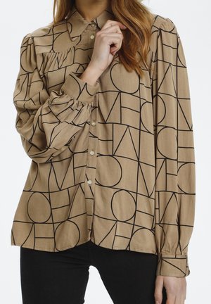 Mujer que lleva una camisa beige con botones, formas geométricas negras, mangas abullonadas y cuello, combinada con pantalones negros.