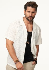 Kurzärmliges, weißes Texturhemd mit dekorativem Muster, braunen Knöpfen, getragen über einem schwarzen Tanktop, ergänzt durch mehrschichtige Halsketten und Armbänder.