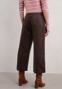 Hnědé kordové kalhoty širokého střihu s dvěma kapsami na zadní straně, doplněné hnědými botami na podpatku. Halenka má růžové a bílé vodorovné pruhy.