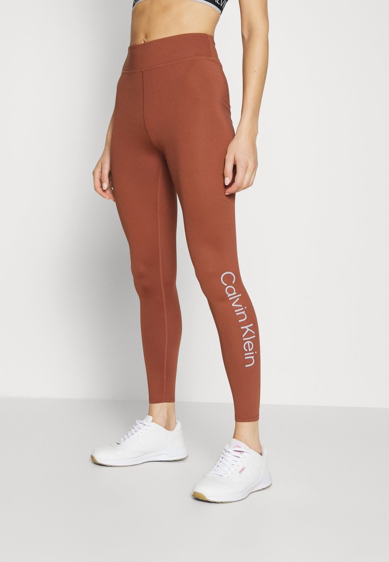 Leggings bruns avec une texture lisse, taille haute, et un logo "Calvin Klein" blanc sur la jambe inférieure. Associés à des baskets blanches.